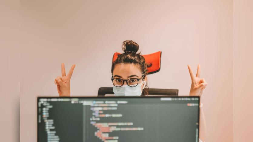 A girl wearing surgical mask behind a computer screen with victory sign in her hands