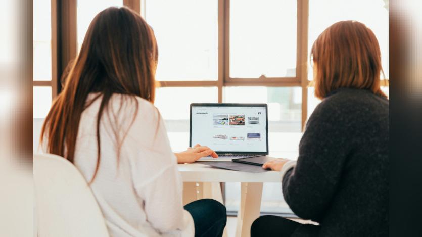 Two women seen from behind, seated on either side of a laptop and facing the same screen.