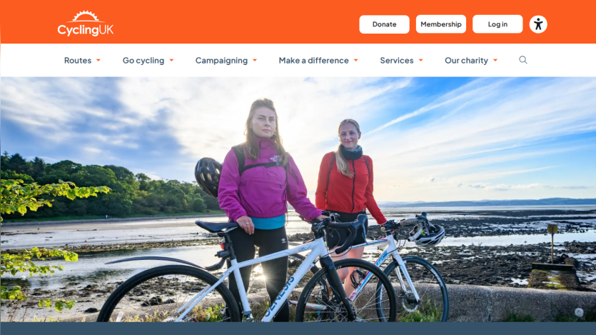 CyclingUK homepage with navigation menu and hero image of two women with bicycles by a coastal landscape