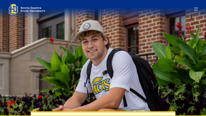 Student in a t-shirt and cap sitting in front of a university building with flowers.
