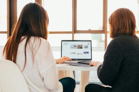 Two women seen from behind, seated on either side of a laptop and facing the same screen.